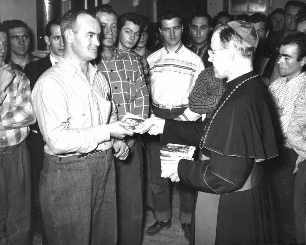 Bishop Couturier hands a document to a worker, surrounded by lumberjacks